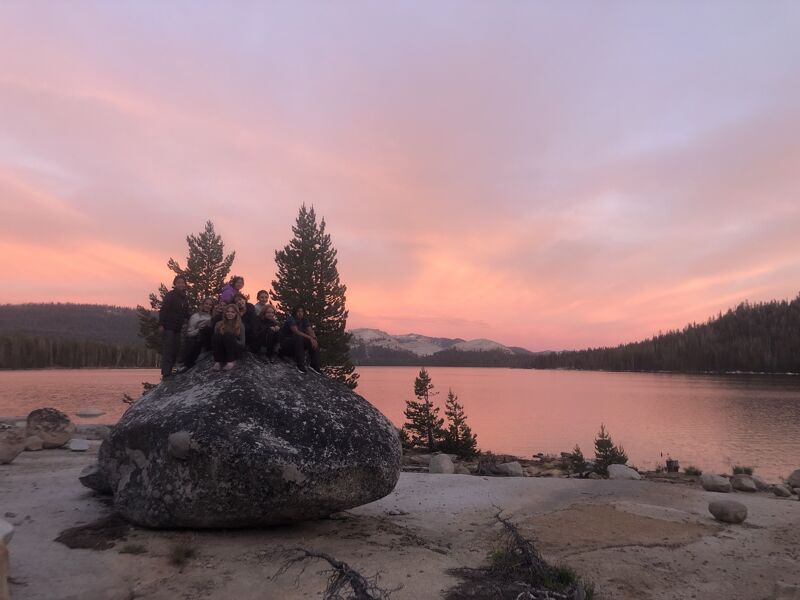 The image shows a serene landscape at sunset. A group of people are gathered on a large rock near a calm lake, with trees silhouetted against the colorful sky. The sky is painted with hues of pink and orange, reflecting on the water's surface. Mountains are visible in the distance, adding depth to the scene.